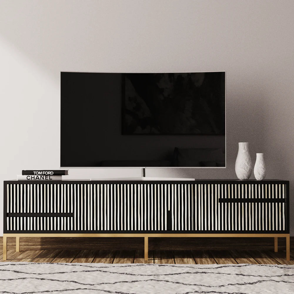 Modern black and gold TV cabinet with vertical slat design, featuring a sleek flat-screen television, designer books, and white decorative vases on top, placed against a neutral wall with a geometric patterned rug below